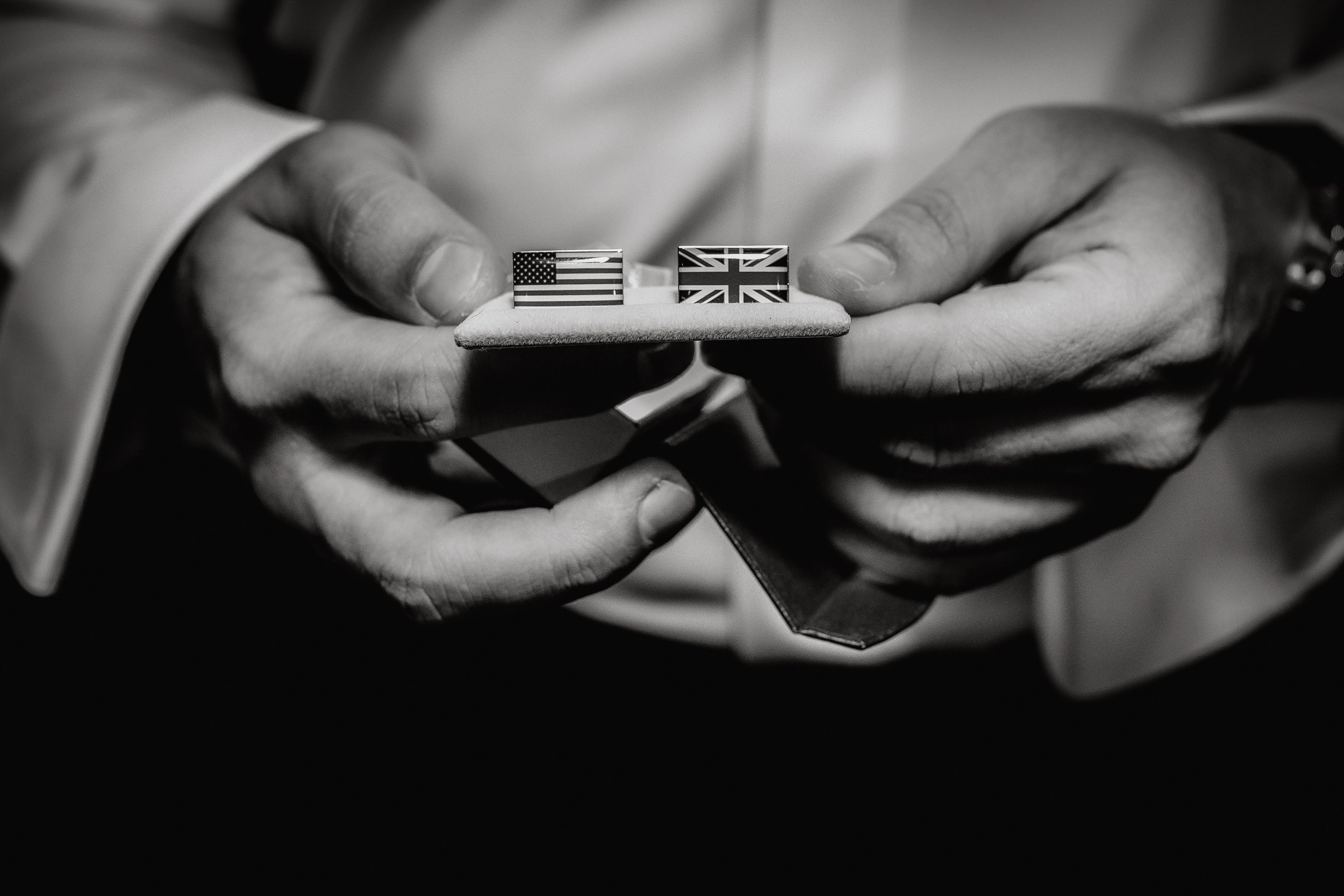 Black and white photo of a person holding a stand with two cufflinks, one with the American flag and the other with the British flag.