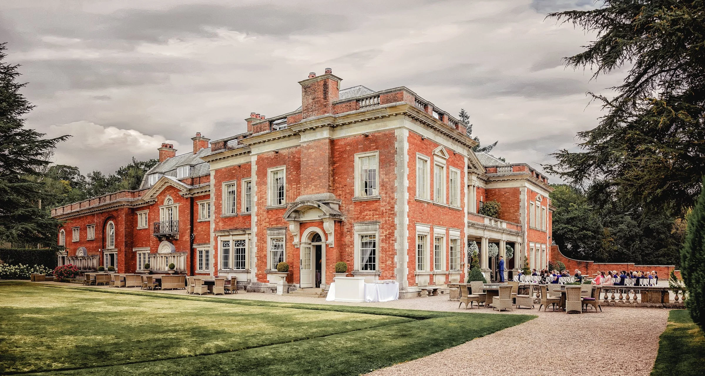 A large, elegant red-brick mansion with white trim and multiple chimneys. Outdoor seating is arranged around the house for a gathering, with some guests seated and others standing, on a cloudy day.