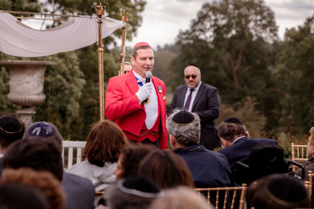 A man dressed in a red jacket with medals, speaking into a microphone at an outdoor event, with audience members wearing yarmulkes and a man in a suit standing behind him.