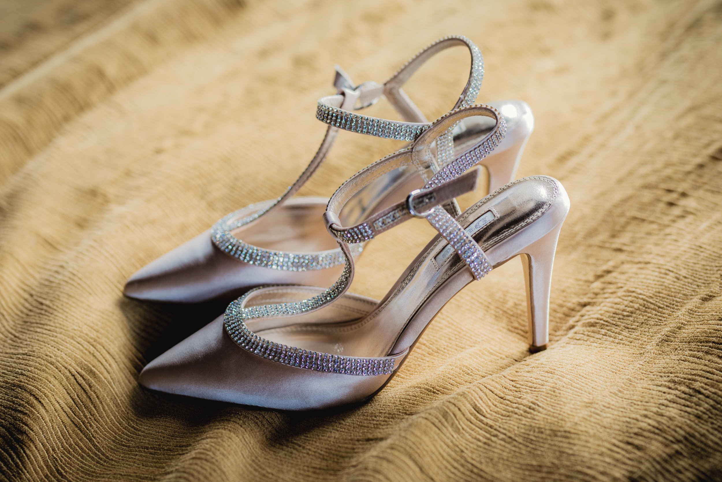 A pair of elegant high-heeled shoes with pointed toes, satin finish, and rhinestone-embellished straps, resting on a textured yellow fabric surface.