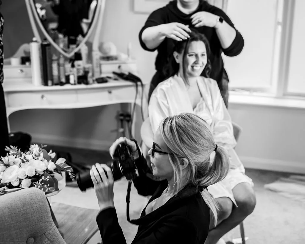 A woman with glasses sitting in a chair, smiling as a hairstylist styles her hair. A photographer with glasses takes pictures of the scene. The room has a vanity with hair products and a mirror, and there is a window letting in natural light.