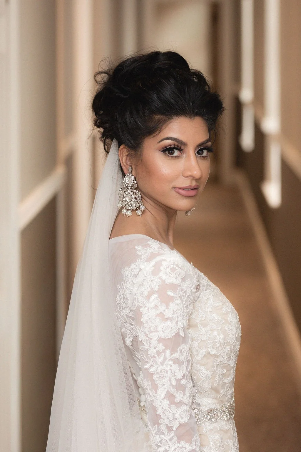 Brunette bride wearing a white lace wedding dress, large statement earrings, and a veil, standing indoors with a hallway background.