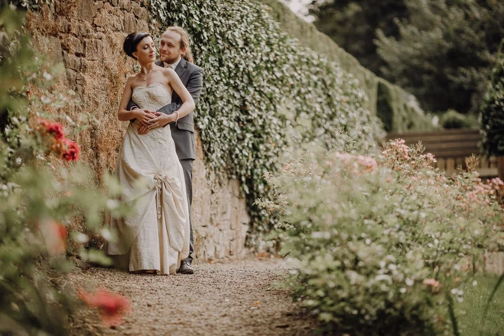 A couple in wedding attire standing by a stone wall, surrounded by pink and white flowers in a garden.