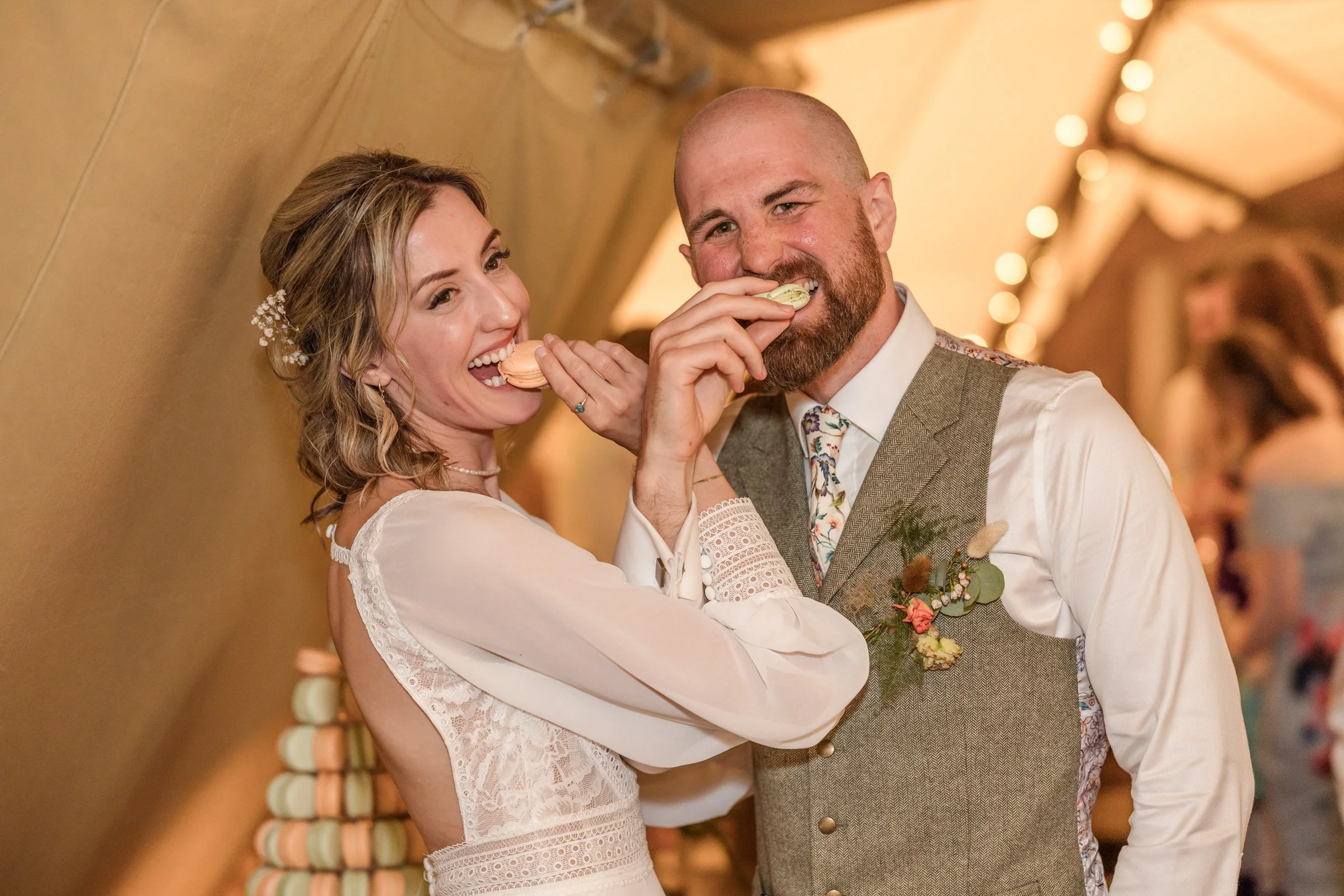 A bride and groom smiling and eating macarons at a wedding reception indoors with warm lighting and string lights in the background.