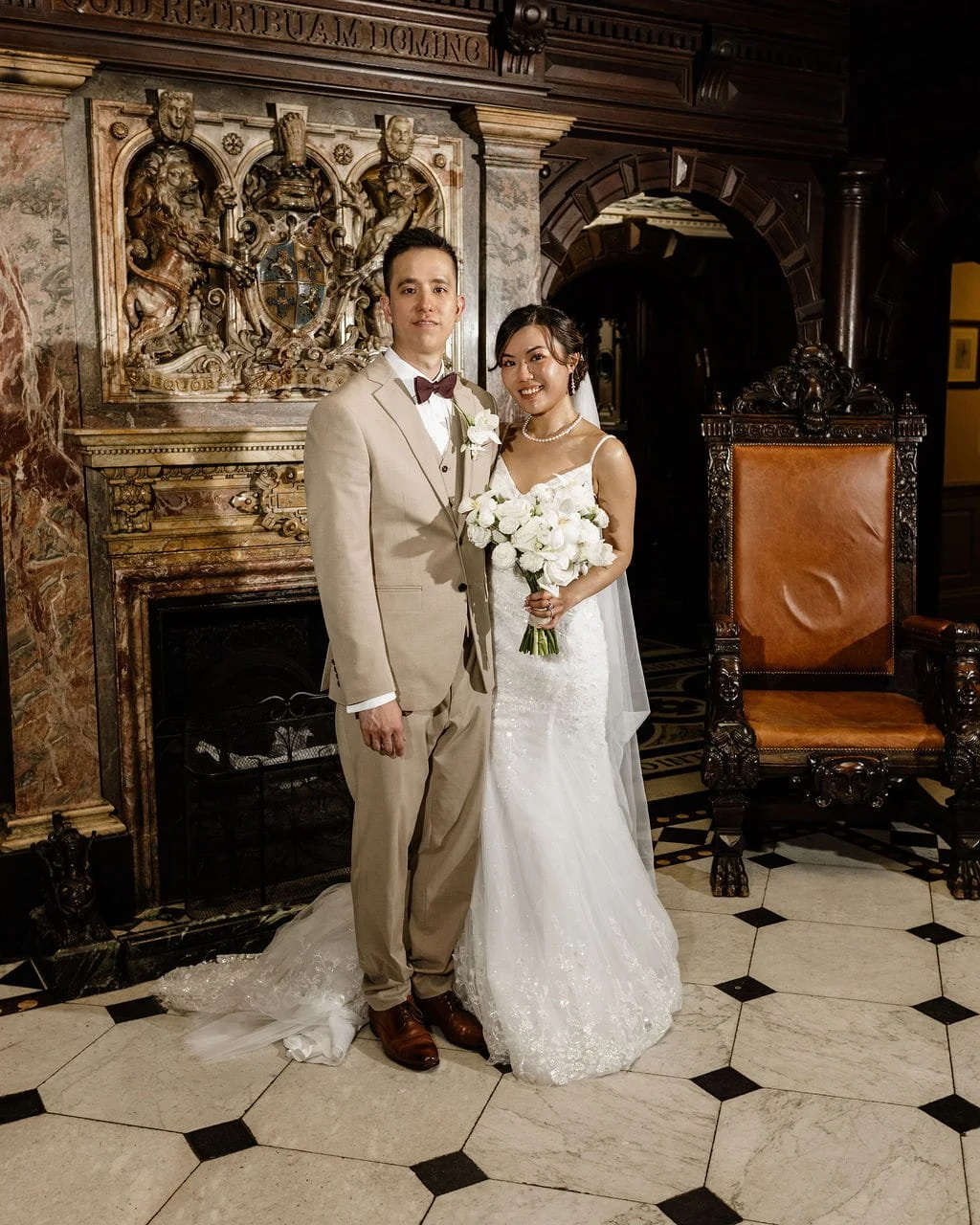 A newlywed couple in wedding attire stands inside a historic, ornately decorated room. The groom wears a beige suit with a bow tie, while the bride wears a white lace wedding gown and holds a bouquet of white roses. They stand in front of a detailed 