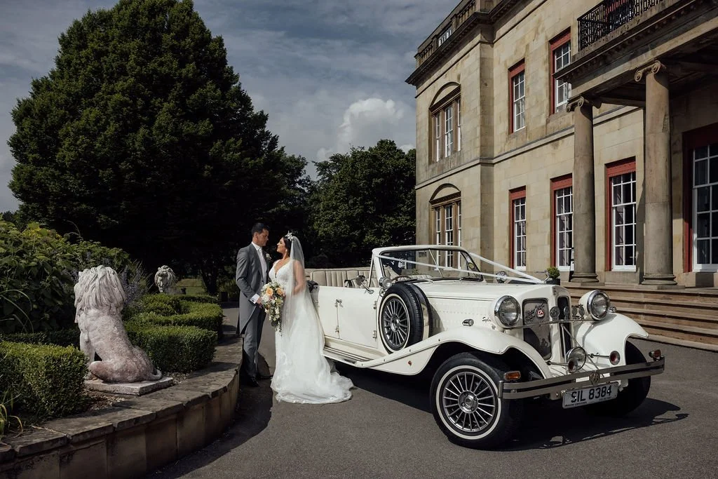 A bride and groom standing next to a vintage white convertible car outside a large elegant building, with lush greenery and lion statues nearby.