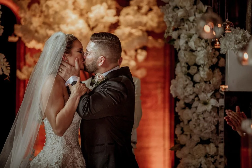 A bride and groom sharing a kiss during their wedding ceremony, surrounded by floral decorations and candles.