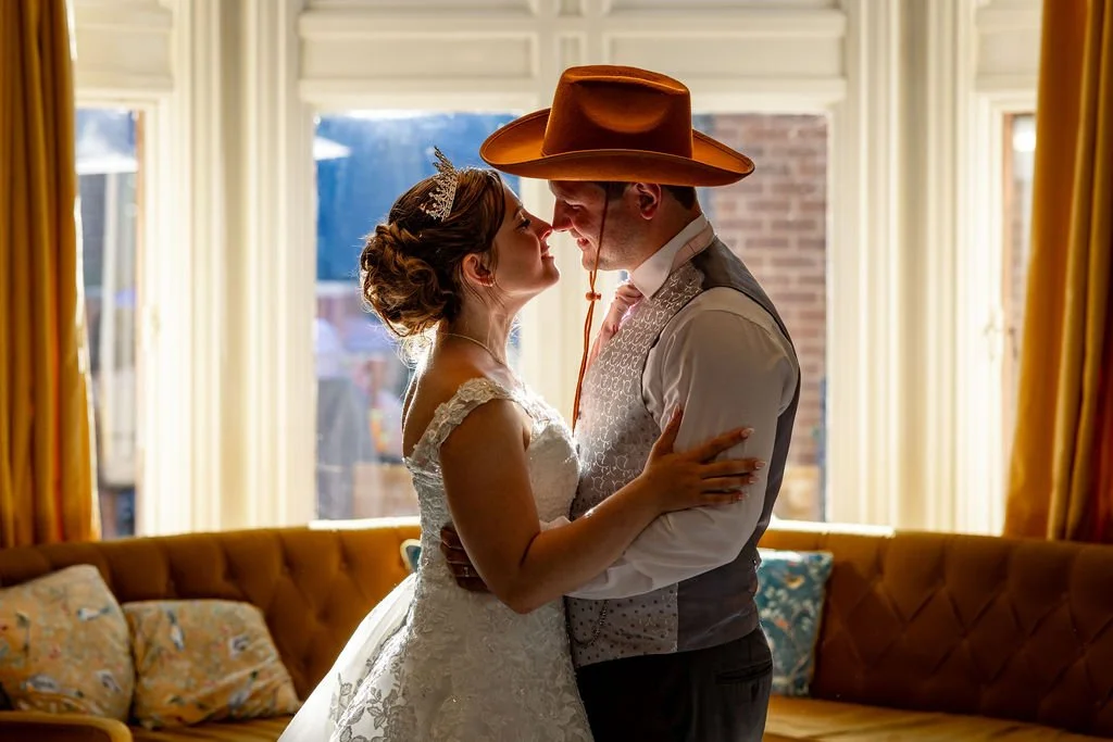 A bride and groom share a close moment indoors, with the bride wearing a white wedding dress and the groom in a cowboy hat and vest, standing in front of windows with yellow curtains.