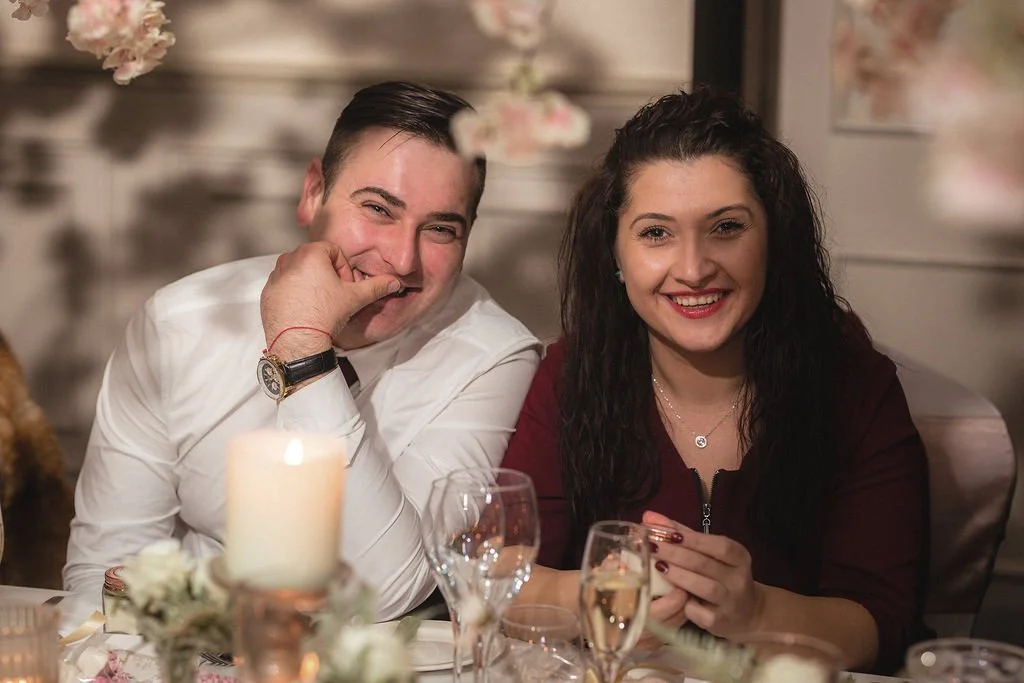 A smiling man with dark hair and a white shirt sitting next to a smiling woman with long dark hair and a maroon top at a dinner table with candles and glasses.