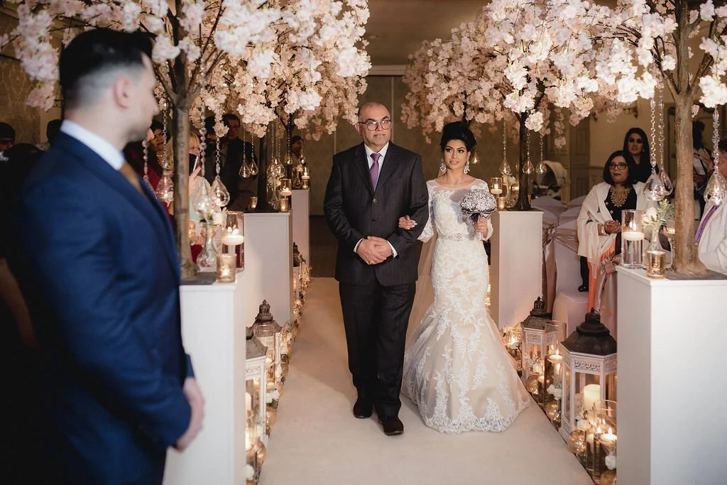 Bride walking down the aisle with her father at her wedding, decorated with pink cherry blossom trees and candlelit lanterns.