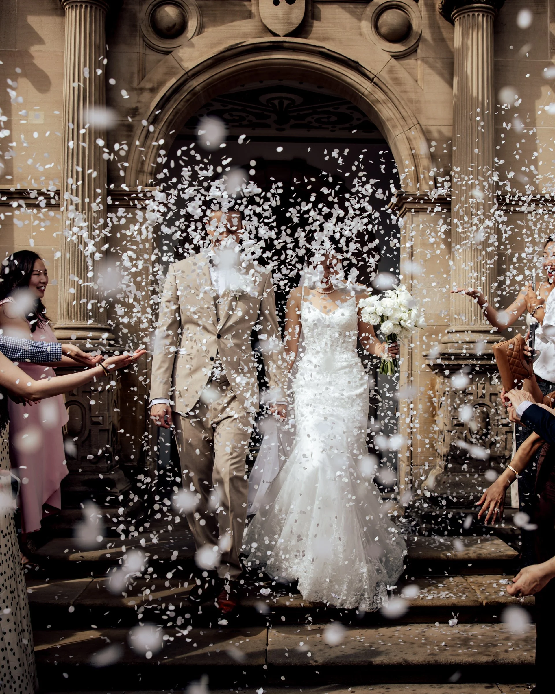 A newly married couple stands in front of a church entrance, surrounded by falling white confetti, with friends celebrating around them. The bride holds a bouquet of white roses, and the groom wears a beige suit, both smiling.