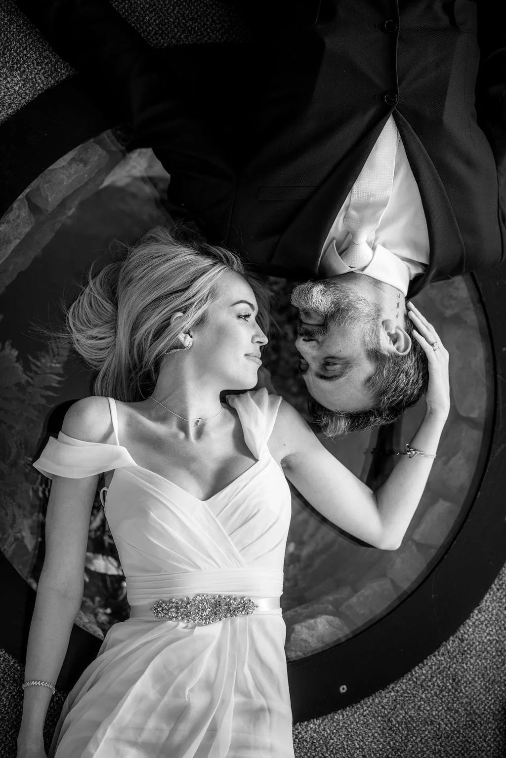 Black and white photo of a bride and groom lying on a round glass surface, faces close, smiling and gazing at each other, woman with long blonde hair in a wedding gown, man with short hair and a beard in a suit, woman touching man's head.