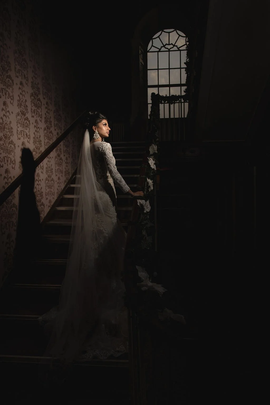 A woman in a wedding dress standing on a staircase next to a decorated floral railing, with dark shadows and a large window in the background.
