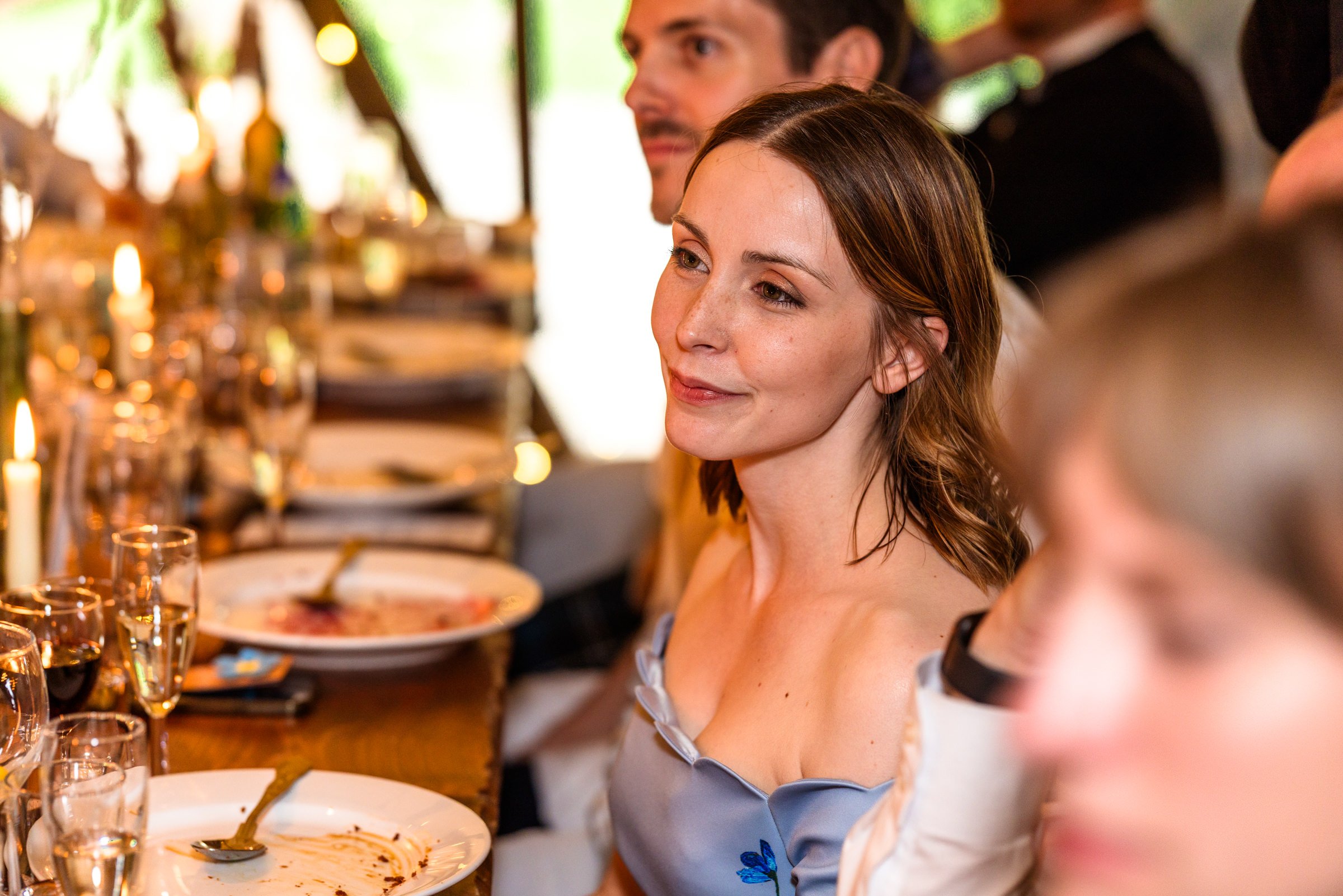 A woman with shoulder-length brown hair and light skin is sitting at a dinner table, smiling softly, surrounded by other people. The table has plates, glasses, and some lit candles.