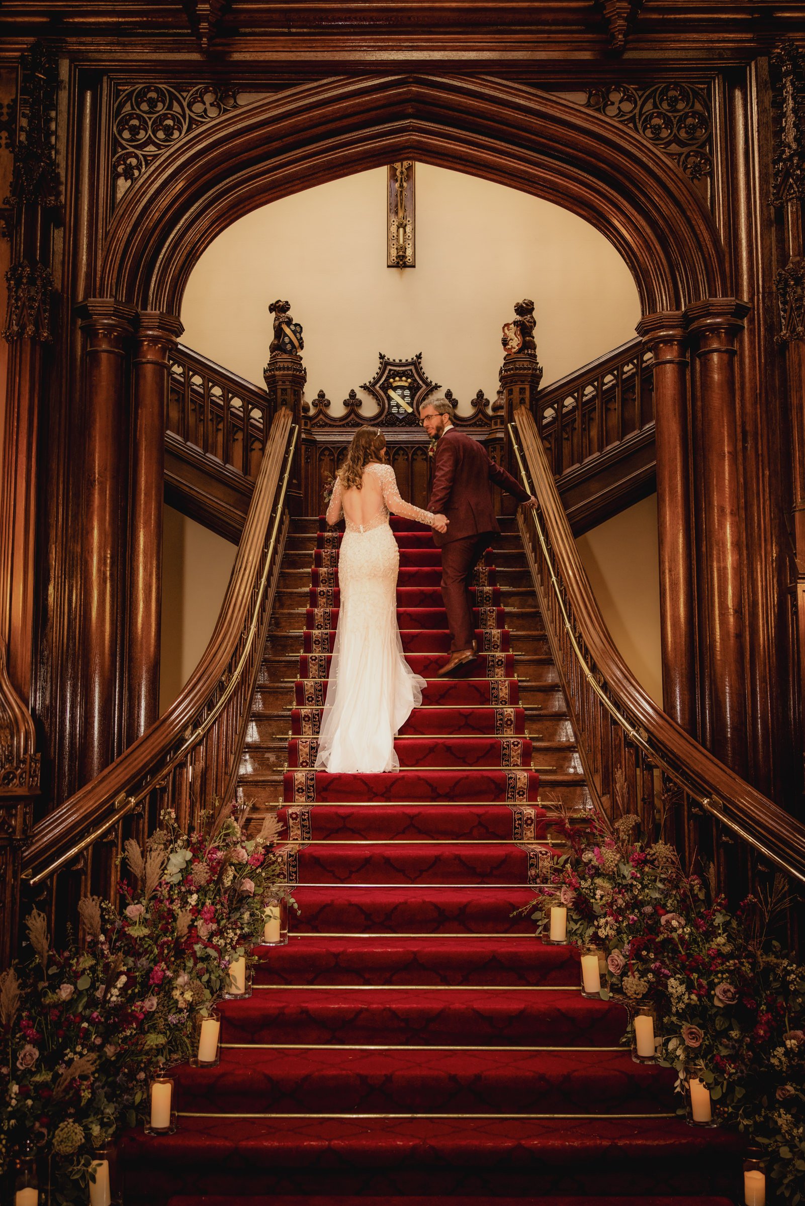 Bride and groom holding hands and ascending a grand staircase with a red carpet, surrounded by floral arrangements and candles, in a historic, ornate wooden interior.