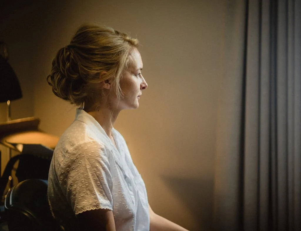 Side profile of a woman with blonde hair styled in an elegant updo, sitting with eyes closed, in a softly lit room with beige walls and curtains.