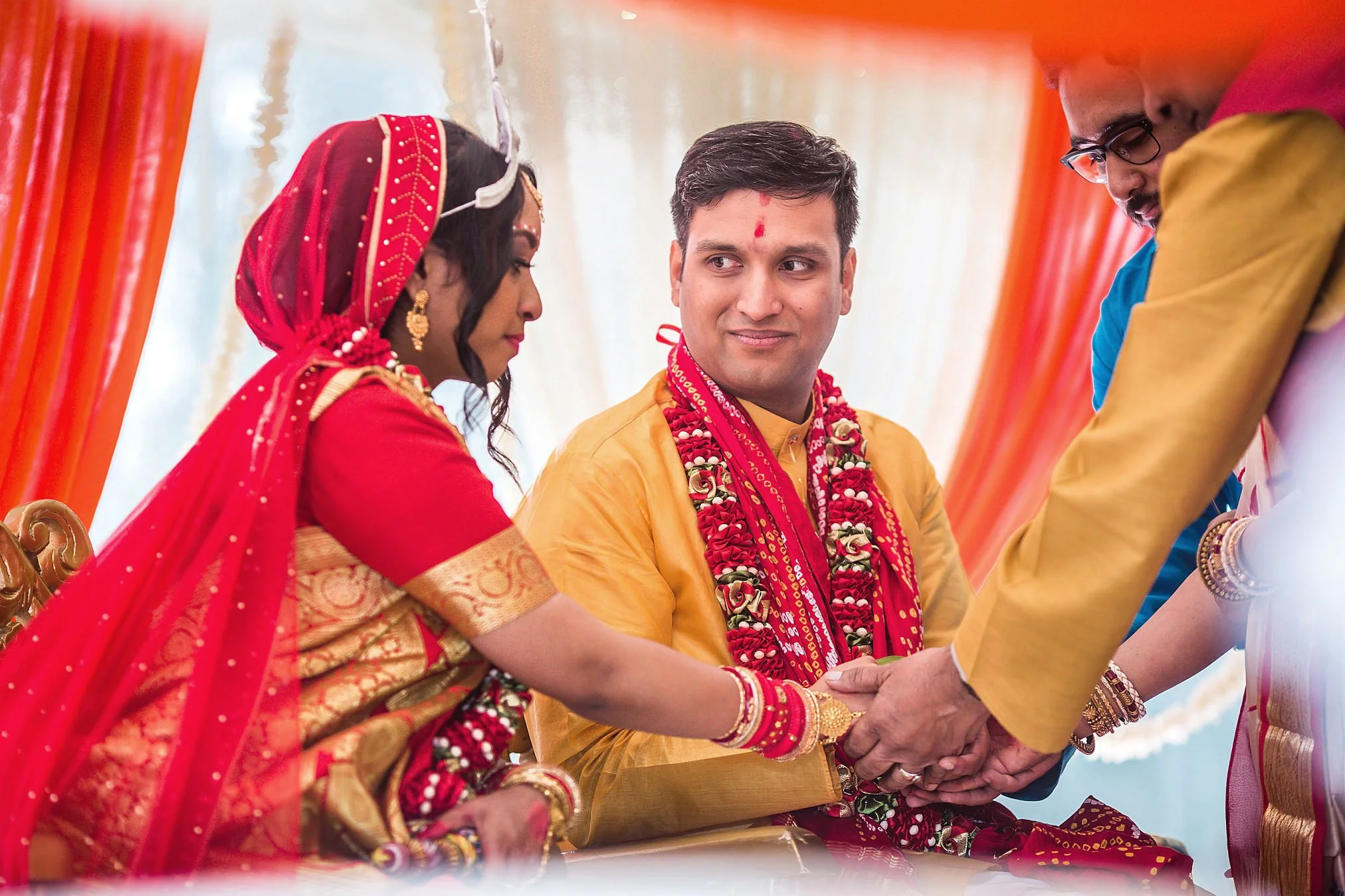 A couple dressed in traditional Indian wedding attire exchanging wedding vows, with two men assisting during the ceremony under an orange and cream decorated canopy.