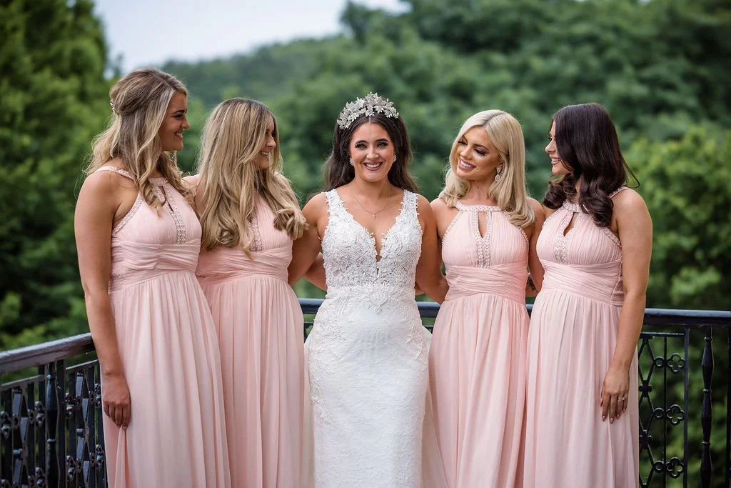 Bride in a white lace wedding gown with a floral crown, surrounded by four bridesmaids in light pink dresses, outdoors on a balcony with greenery in the background.