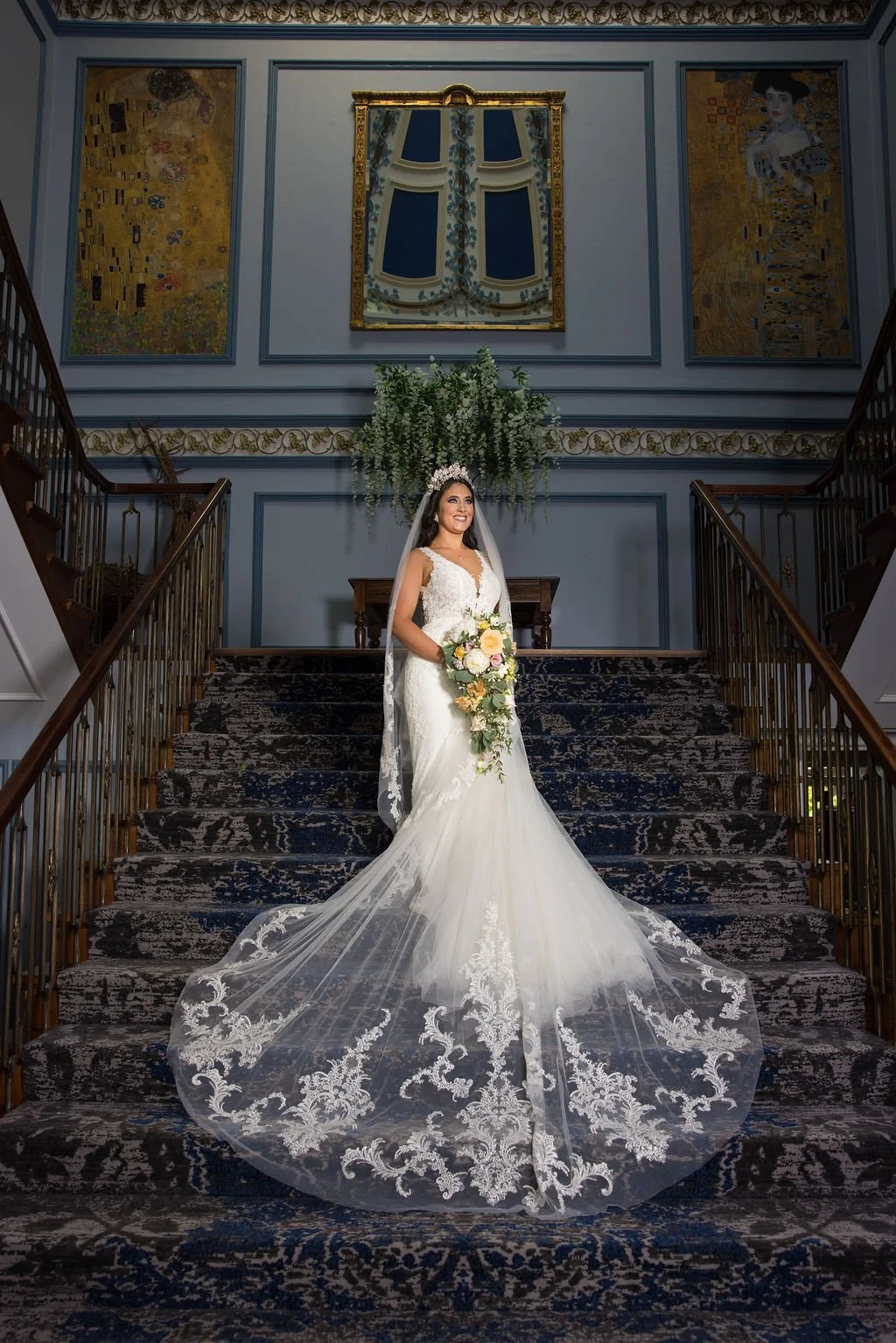 Bride in white wedding gown holding a bouquet, standing on a staircase in an ornate interior with blue walls, gold accents, and artwork, with a floral arrangement behind her.