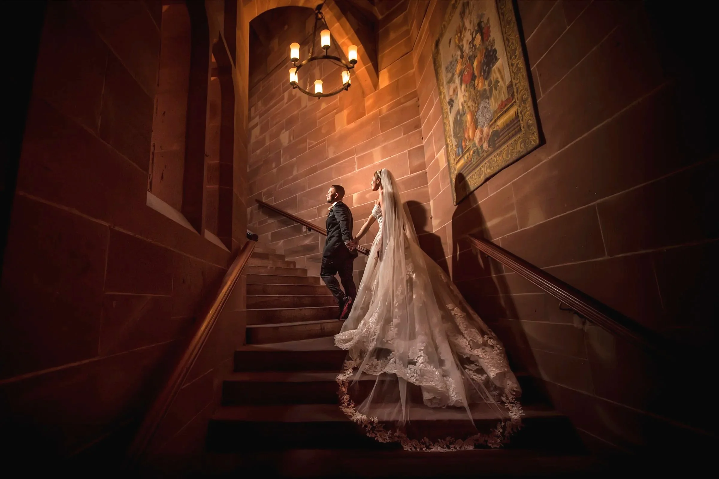 A bride and groom holding hands as they walk up a wooden staircase in a dimly lit, elegant interior with brick walls and a large chandelier.