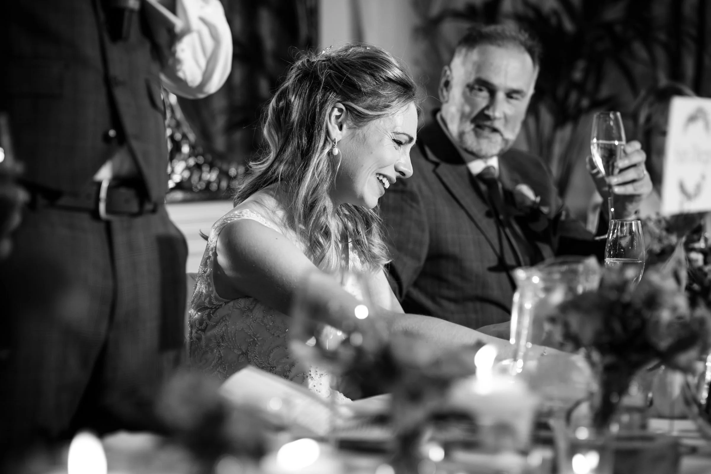 A woman laughing and smiling at a formal event, sitting at a table with a man, in black and white.