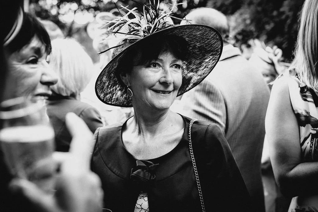 A smiling woman wearing a large, wide-brimmed hat decorated with flowers at a social gathering.