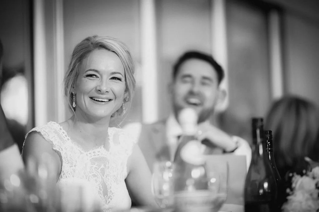 A smiling woman with blonde hair, wearing a lace dress, sitting at a table during a social gathering. A man in the background is laughing, and other people are partially visible at the table.