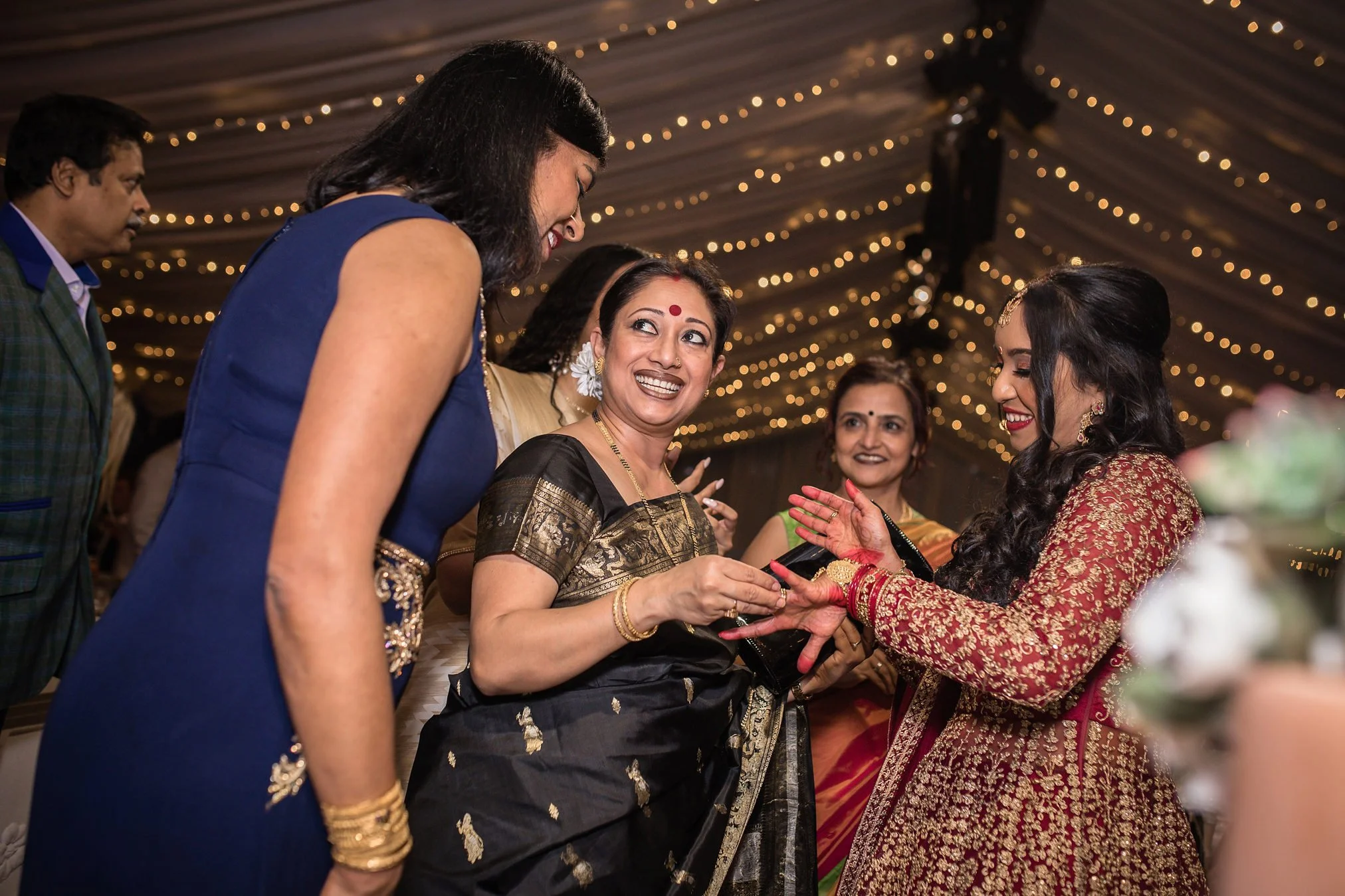 Asian bride in red and gold wedding dress shows off her wedding ring to female guests inside a marquee with golden lights above them.
