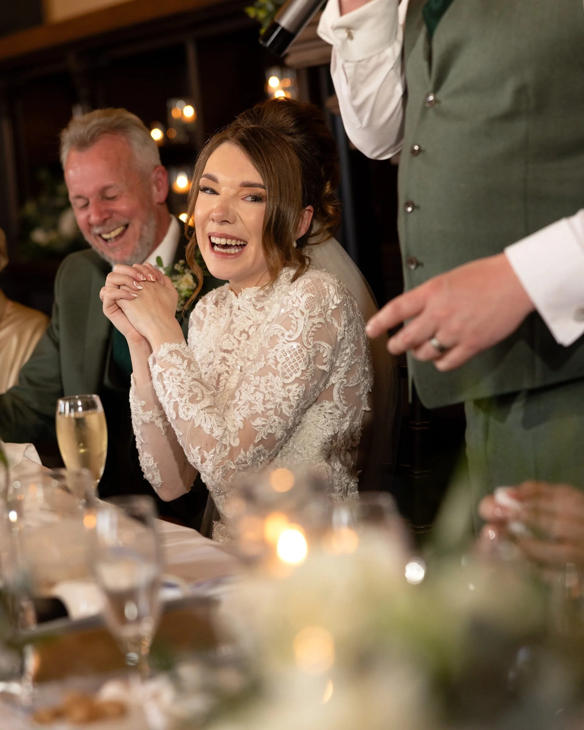 A bride in a lace wedding dress sitting at a table during a wedding celebration, smiling with clasped hands. An older man in a green suit is sitting beside her, also smiling. A person wearing a green vest and white shirt is standing nearby, holding a