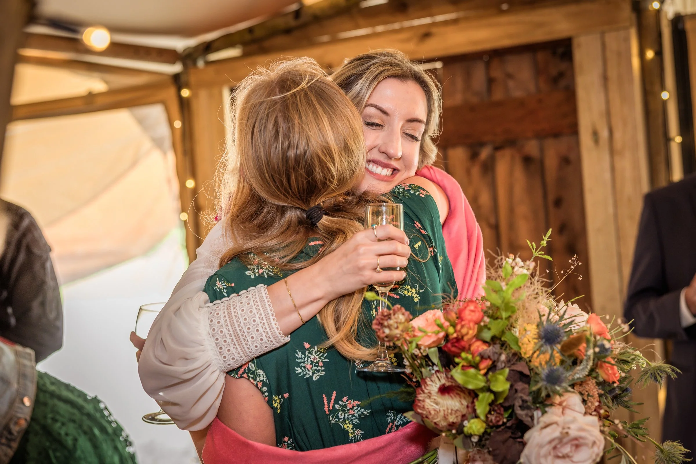Two women hugging at a celebration, one holding a glass of champagne and a large floral bouquet, smiling warmly in a rustic indoor setting.