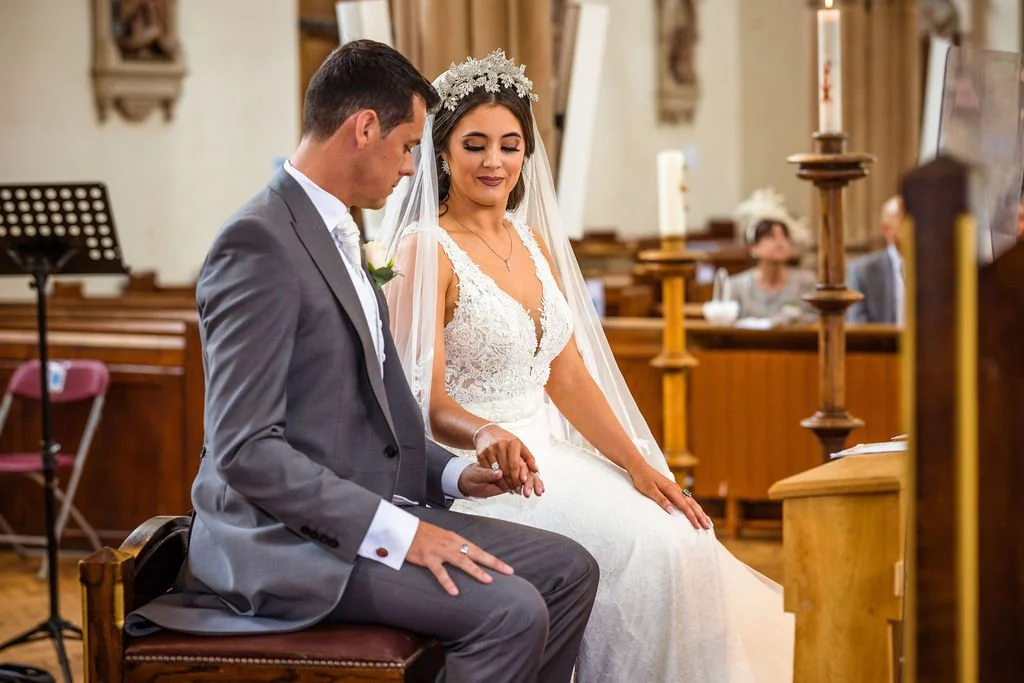 A bride and groom seated hand in hand during their wedding ceremony inside a church.