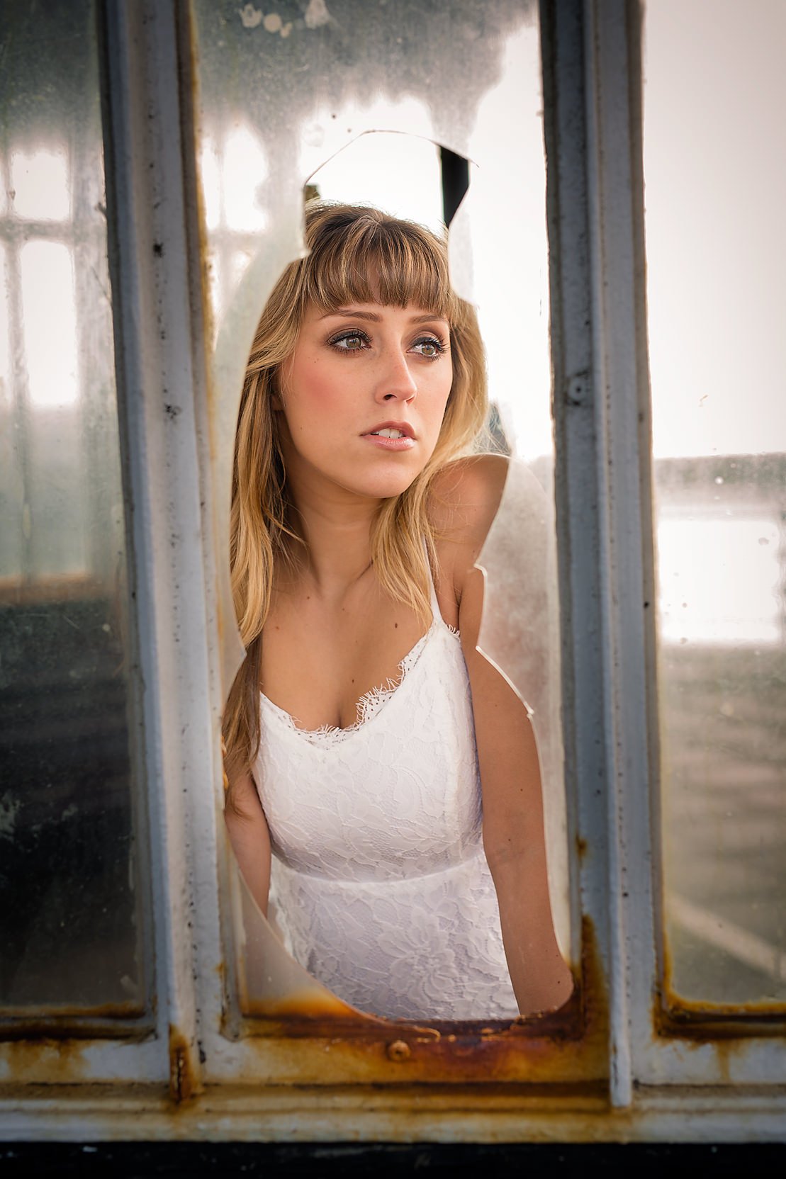 A young woman with blonde hair and blue eyes looking through a rusty, broken window. She is wearing a white lace camisole and appears contemplative or concerned.