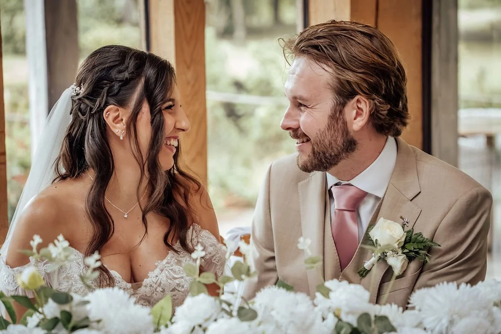 A bride and groom smiling at each other during their wedding ceremony, with floral decor in the foreground.