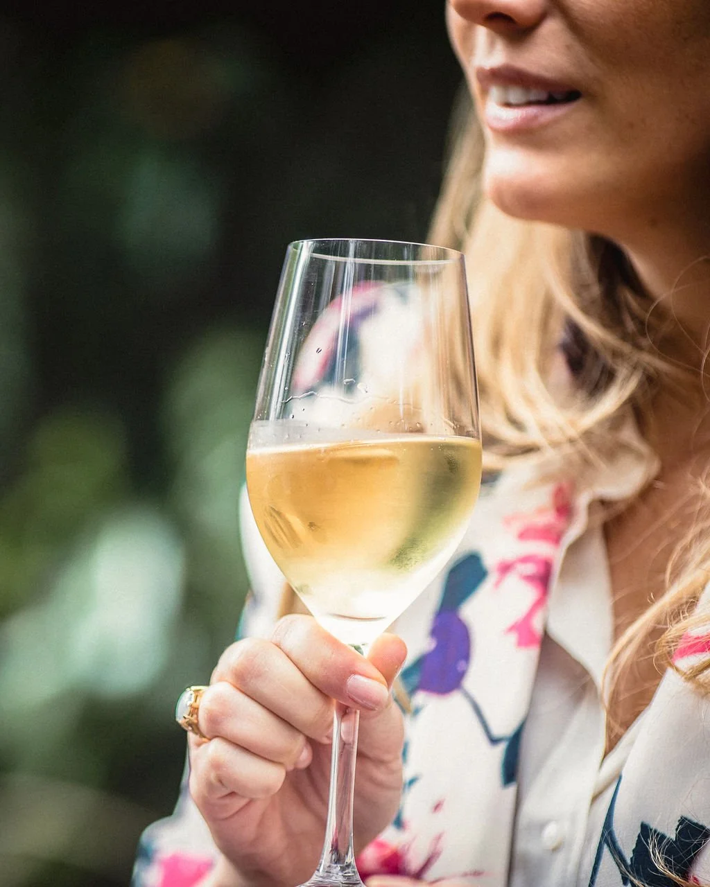 Close-up of a woman holding a glass of white wine, with part of her face and floral-patterned blouse visible.
