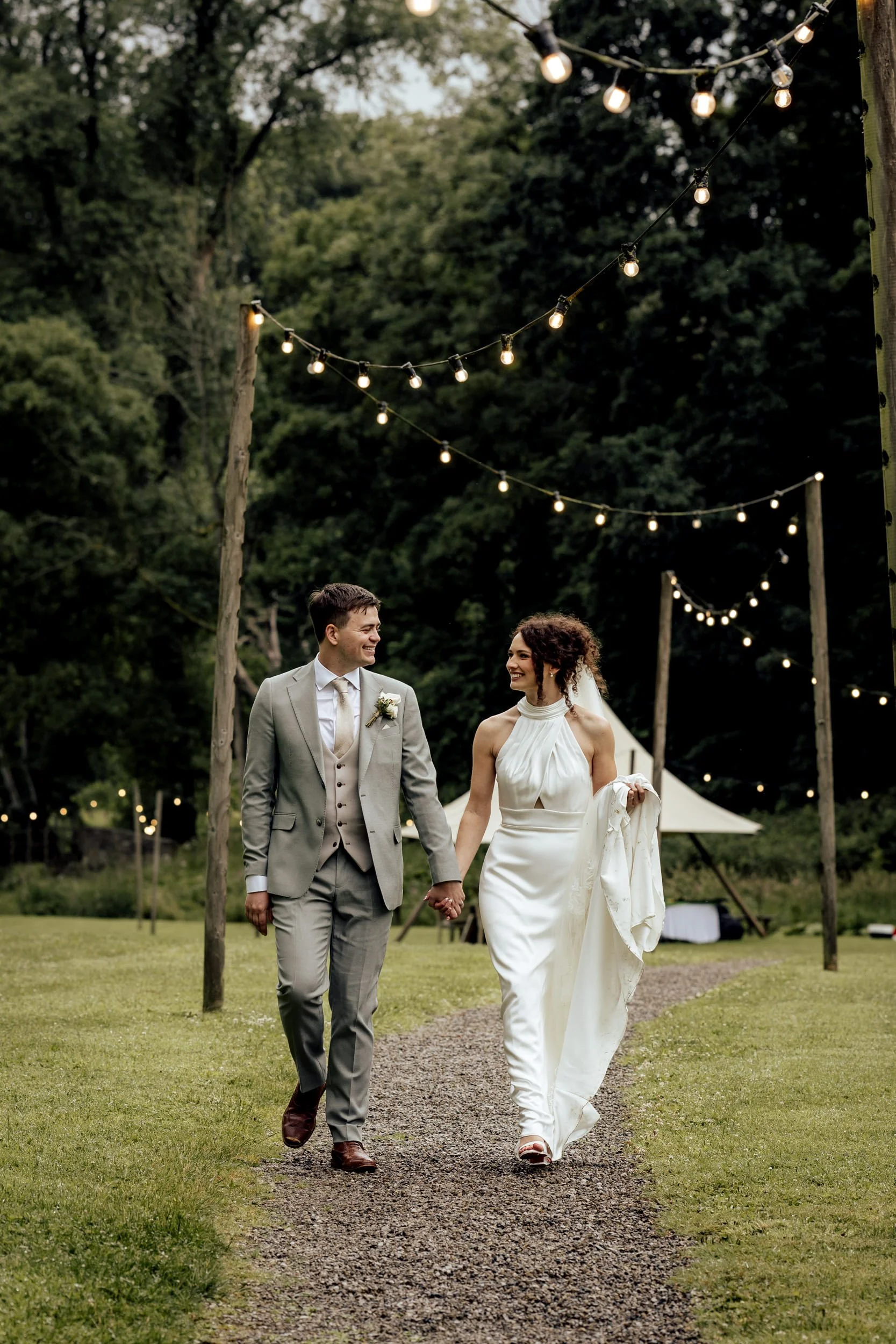 A newlywed couple holding hands and walking outdoors on a gravel path, smiling at each other, under string lights in a lush green setting.