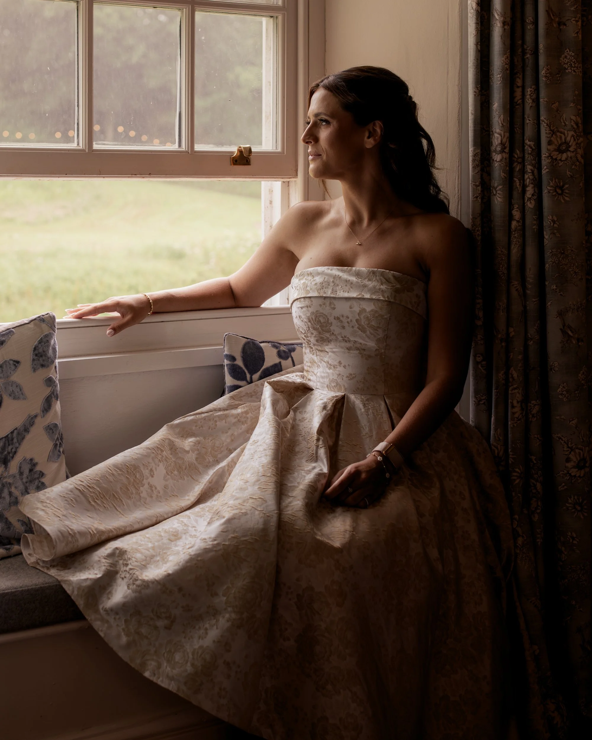 A woman with dark hair styled in loose waves, wearing a strapless cream-colored dress with a floral pattern, sitting on a cushioned window seat and looking out of a window. The window has multiple panes, and there are curtains with a floral design on