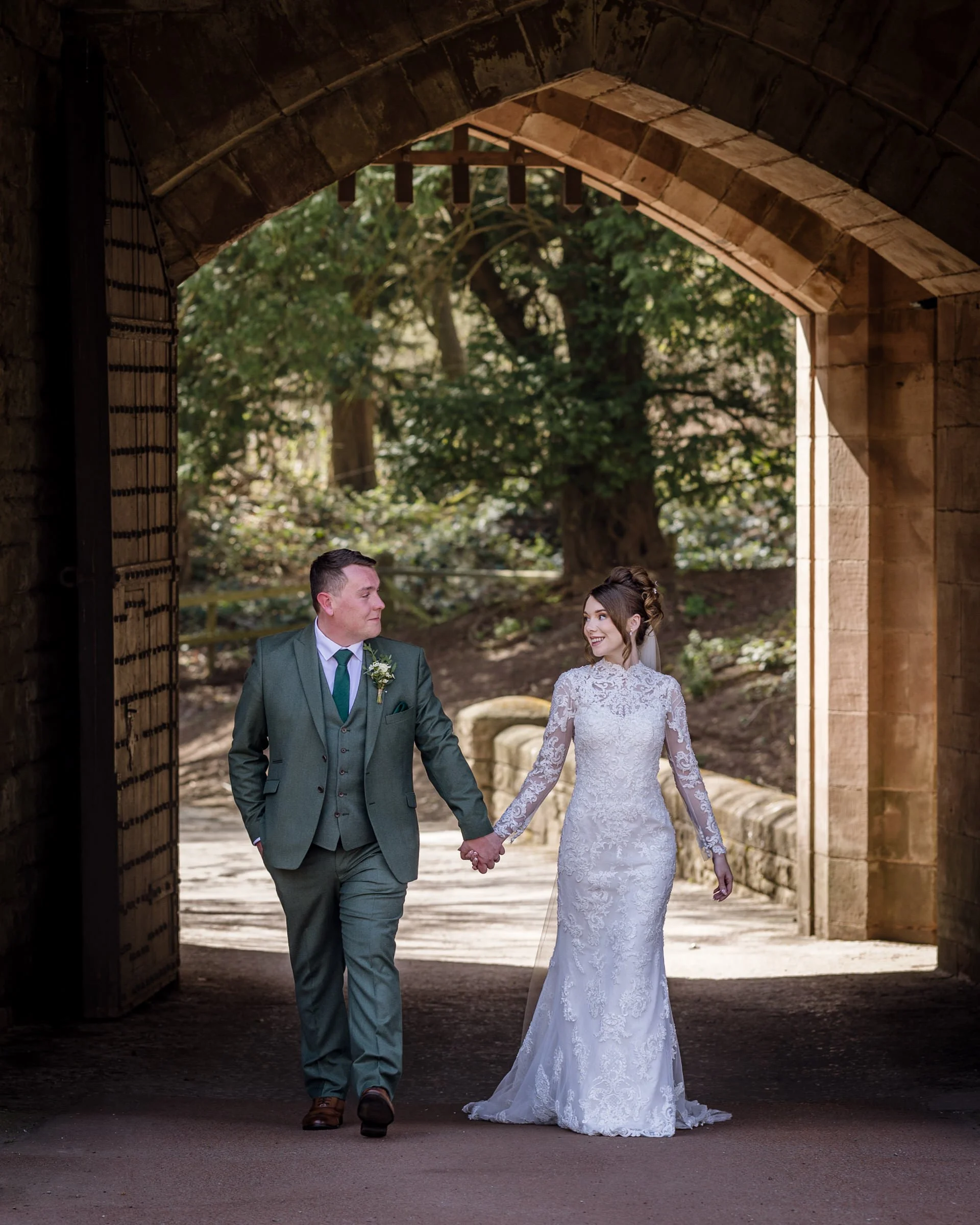 A bride and groom walking hand in hand under an arched stone gateway, with greenery in the background. The bride is in a white lace wedding dress, and the groom is in a green suit with a boutonnière.