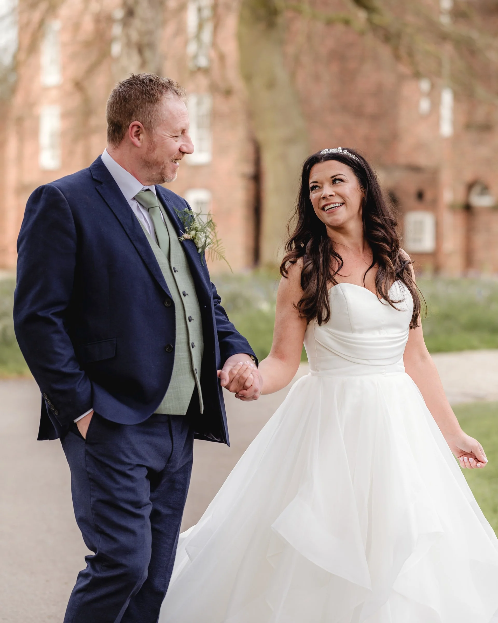 A bride and groom holding hands and smiling at each other outdoors during their wedding, with a brick building and trees in the background.