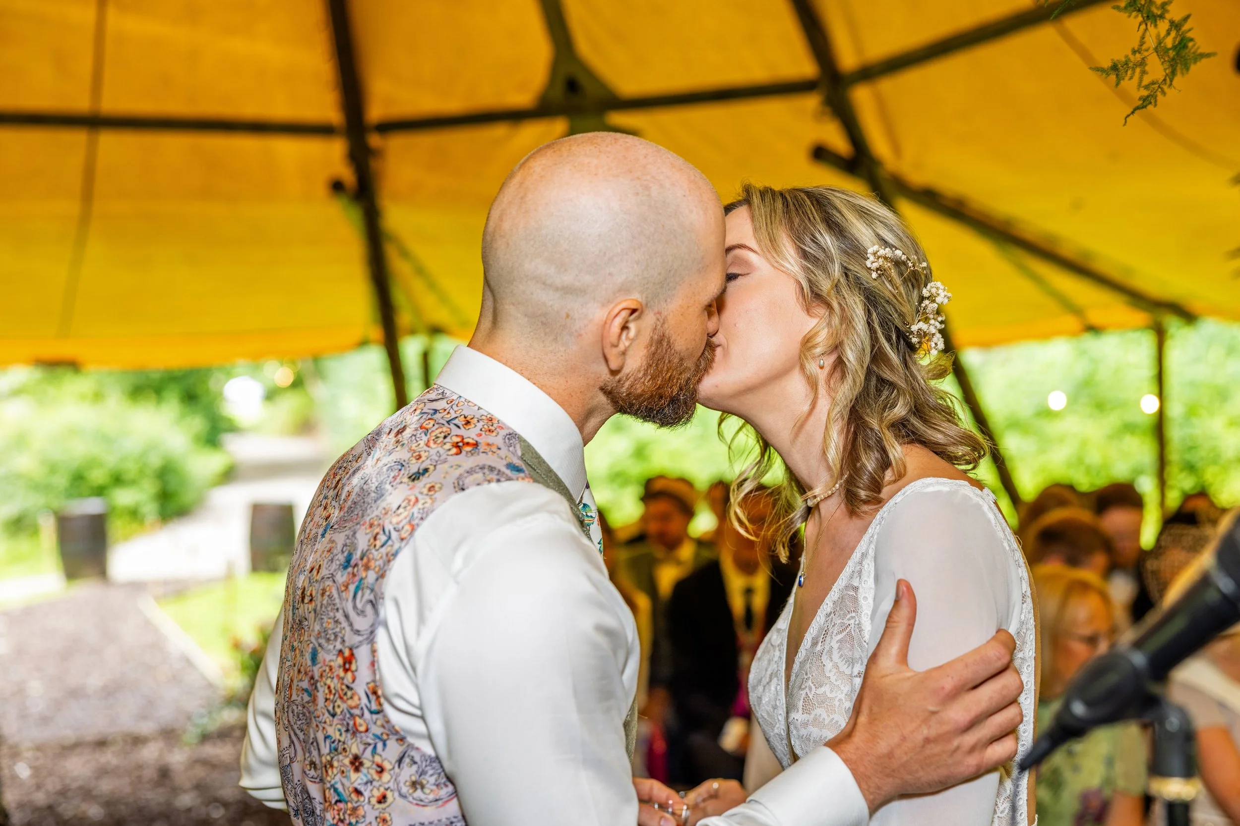 A bride and groom kiss during their wedding ceremony under a yellow canopy, with guests seated behind them.