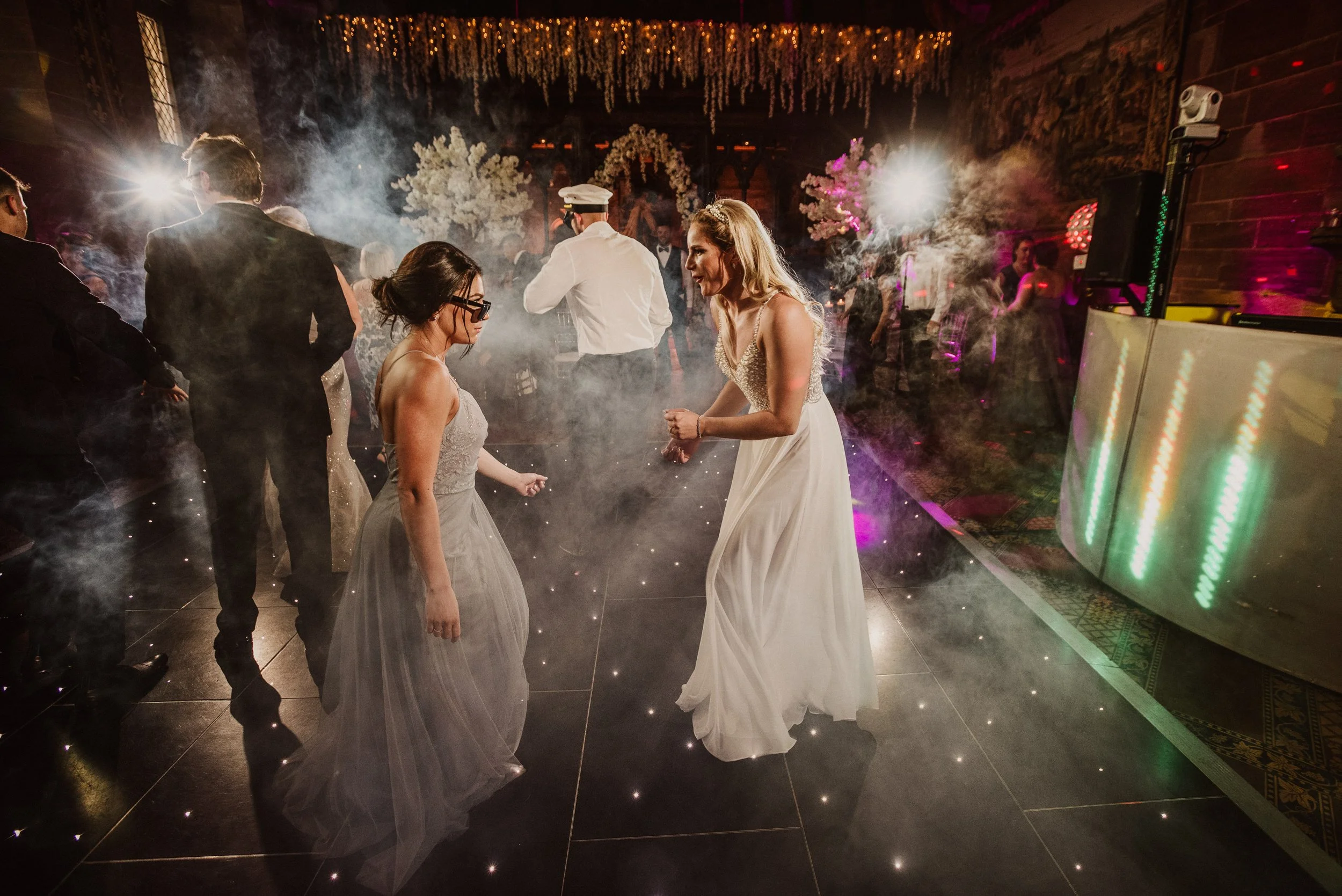 People dancing at a wedding reception in a decorated indoor venue with purple and pink lighting, fog effects, and a DJ booth.