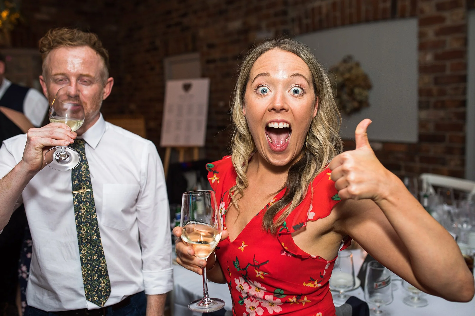 A woman in a red floral dress with an excited expression, giving a thumbs-up, holding a glass of white wine, at a celebration or party.