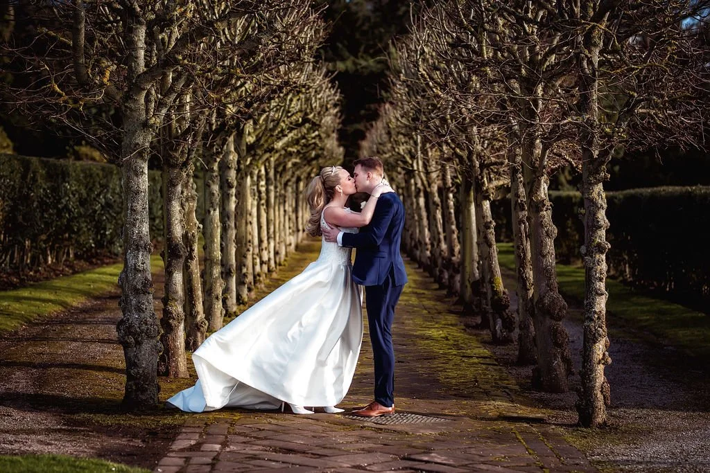A bride and groom share a kiss in a tree-lined pathway, with the bride in a white gown and the groom in a navy suit.