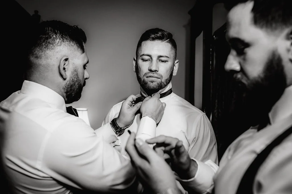 Three men getting ready, one helping the other adjust a bow tie in black and white photo.