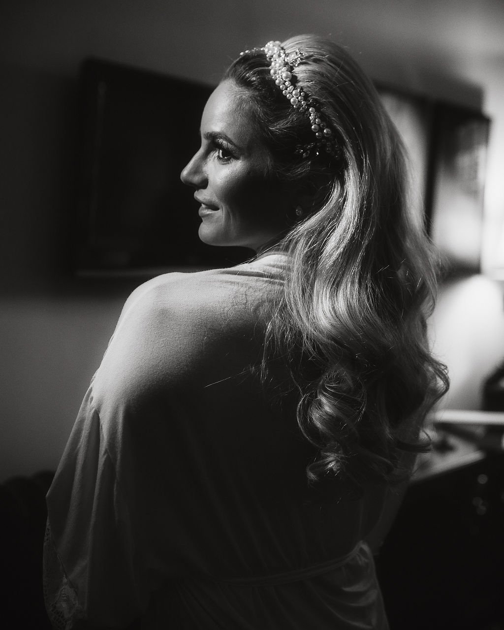 Black and white photo of a woman with wavy hair, wearing a pearl hair accessory, turned to the side with a soft smile, in an indoor setting.