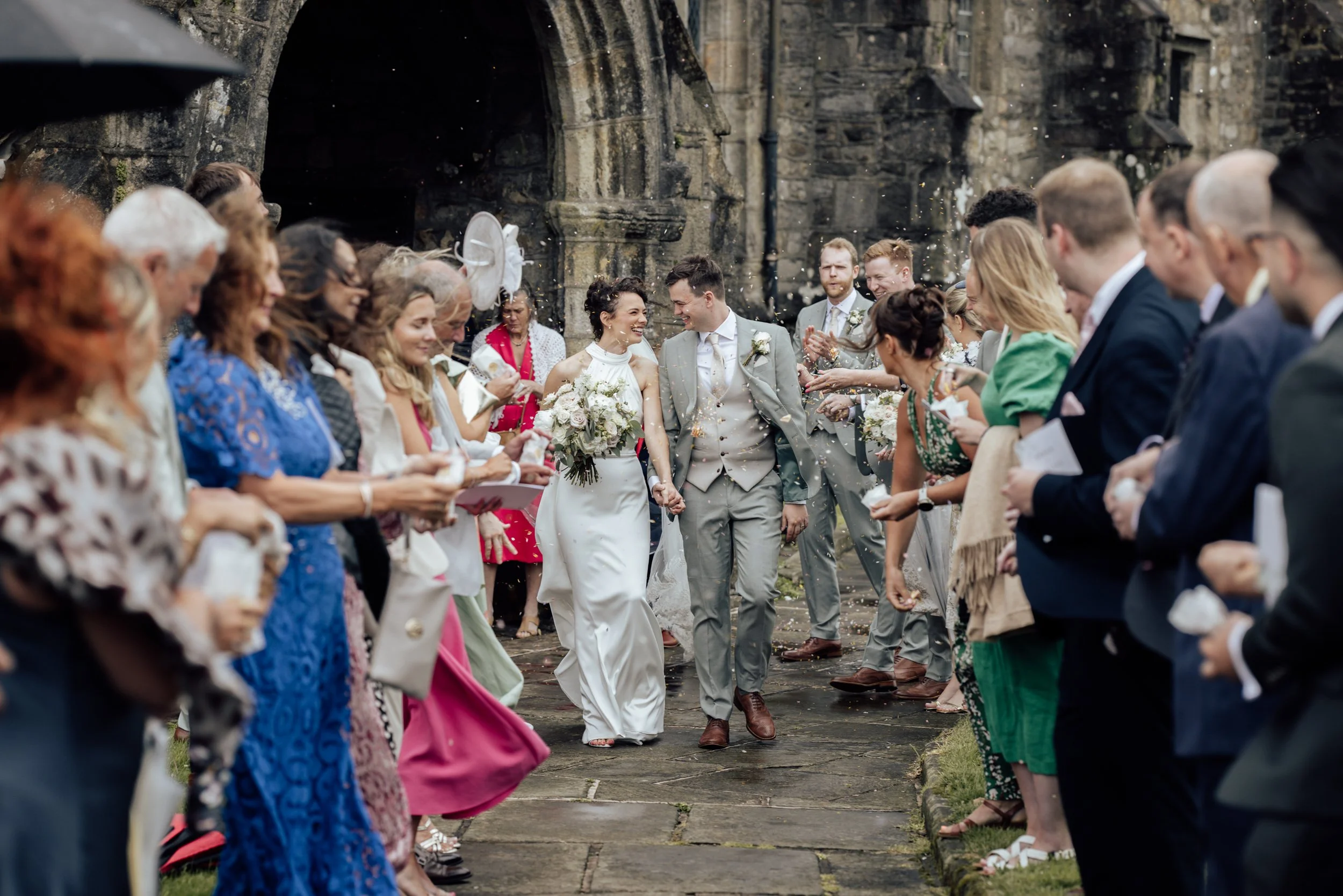 A bride and groom hold hands and walk through a line of wedding guests outside a stone church, celebrating their wedding with confetti and smiles.