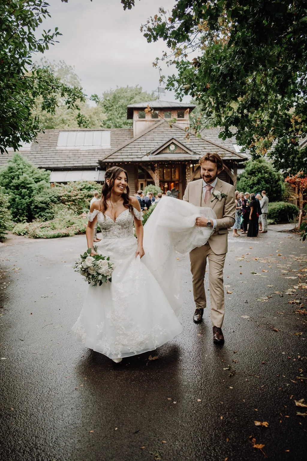A bride and groom are walking outside a rustic wedding venue, smiling and holding wedding attire, with guests in the background and lush green trees surrounding them.