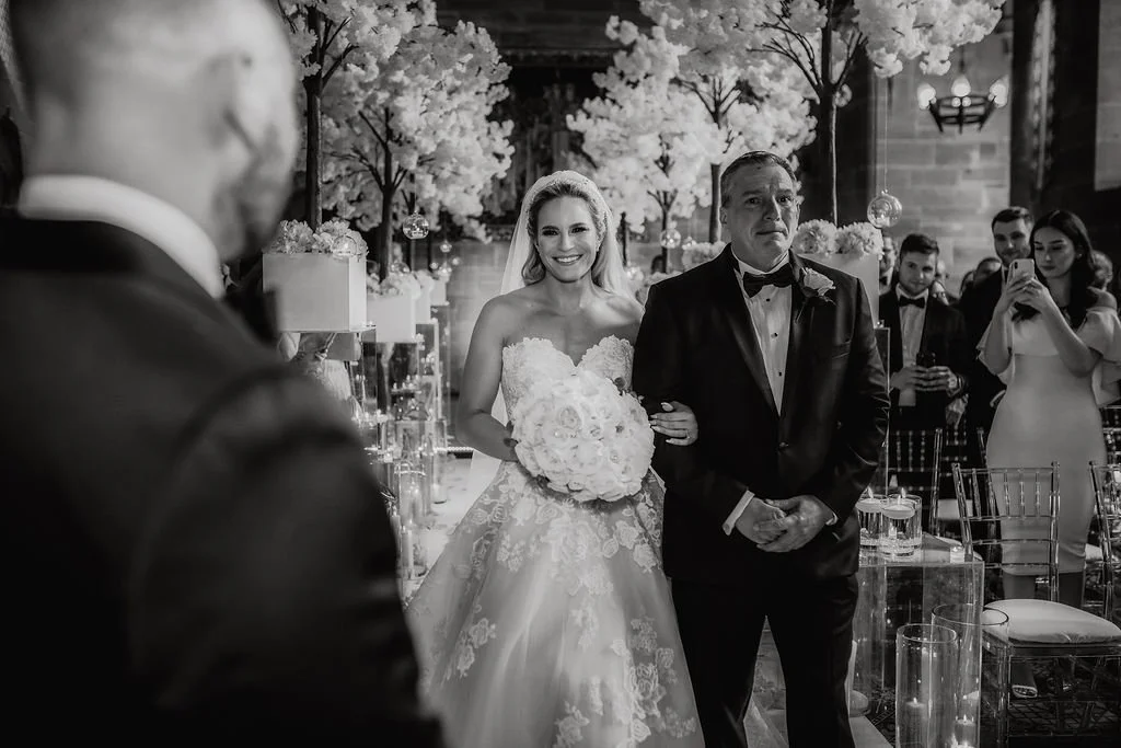 Bride in wedding gown holding bouquet walking down aisle with father during wedding ceremony, guests watching in background, decorated with flowers and floral arrangements.