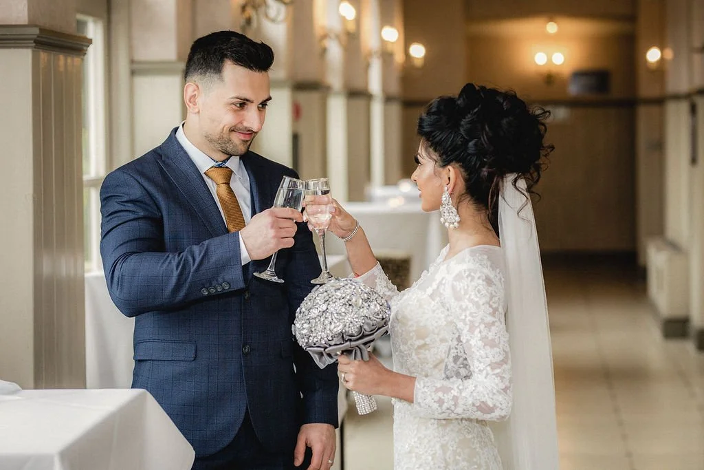 Bride and groom at wedding toast, dressed in wedding attire, holding champagne glasses, in a decorated venue.