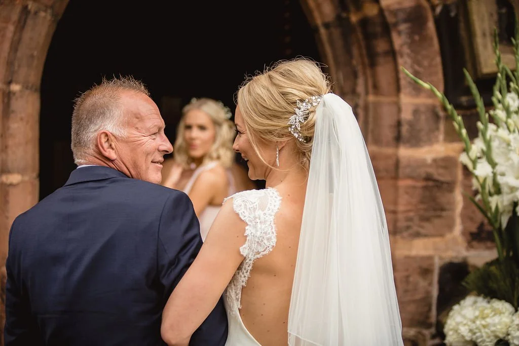 A bride and an older man, possibly her father, sharing a joyful moment at a wedding ceremony, with a woman in the background and floral arrangements visible.