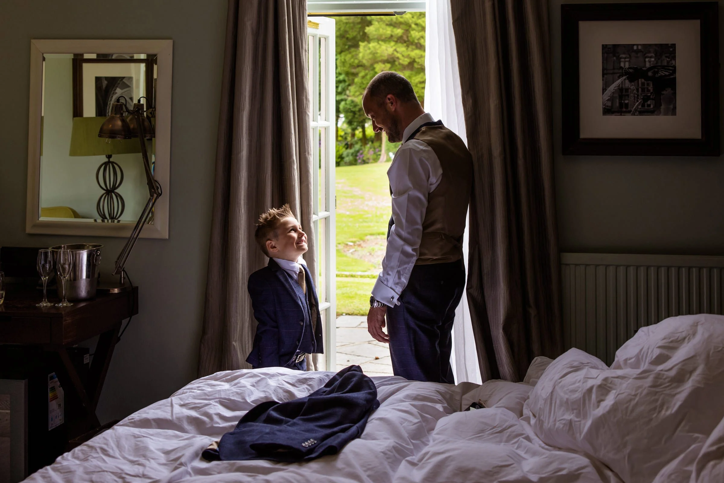 Groom and father looks lovingly at his young son in the open doorway of his bedroom at Mottram Hall.